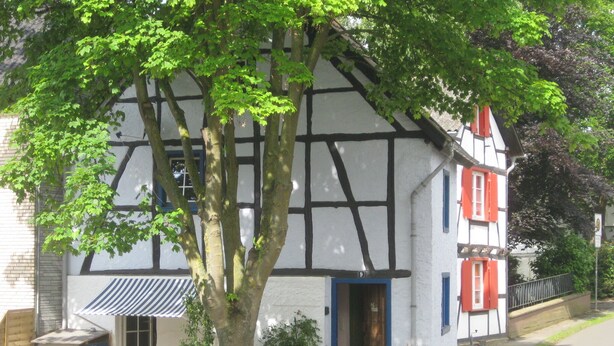 Ein traditionelles Fachwerkhäuschen mit sichtbarer Tür und Fenstern, umgeben von einem Holzzaun und einem Baum.