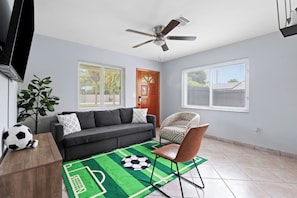 A living room with a grey sofa, a patterned armchair, a green sports-themed rug, and a ceiling fan.