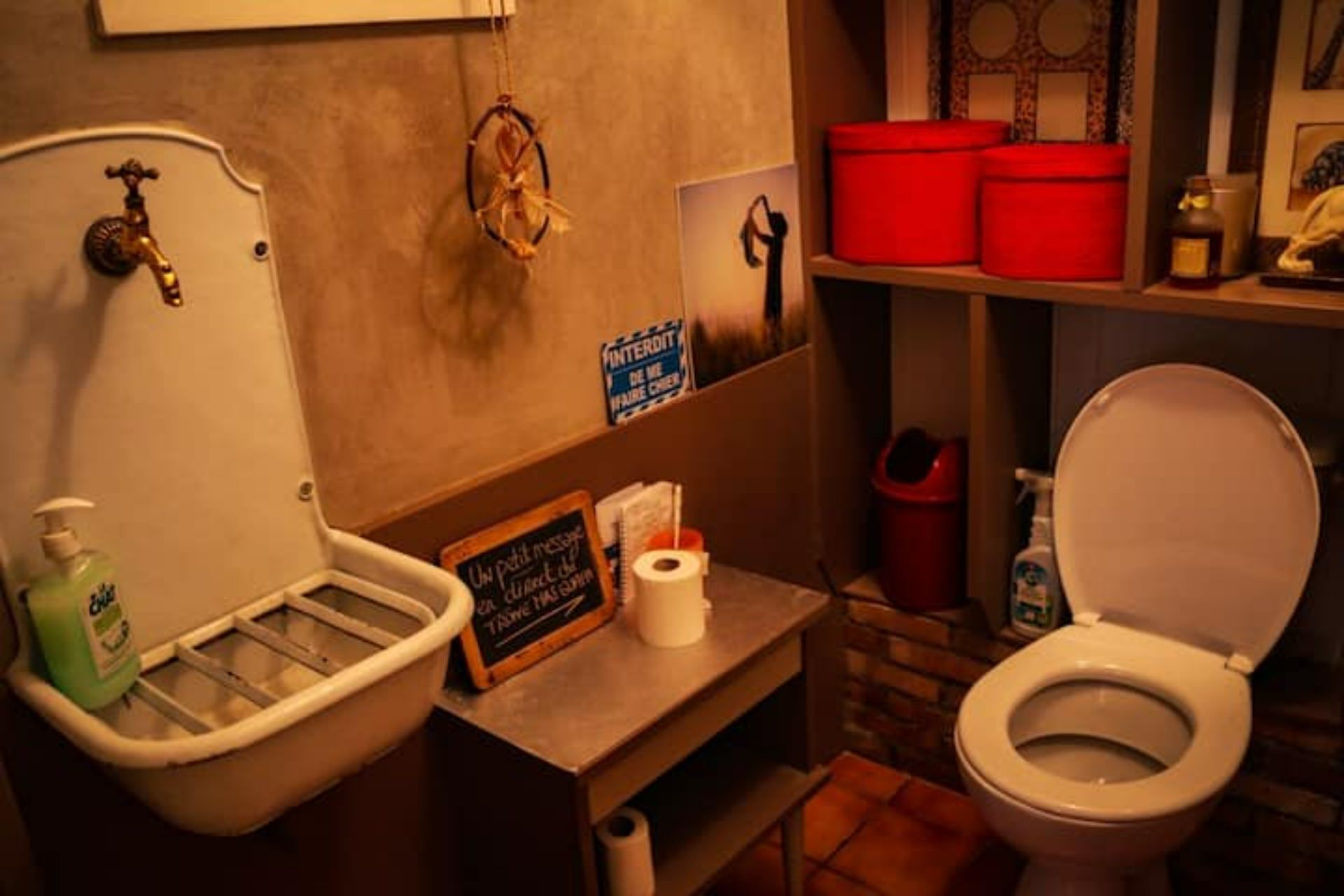 Une salle de bain avec un lavabo blanc, des toilettes, une étagère avec des récipients rouges et un tableau noir avec des inscriptions.