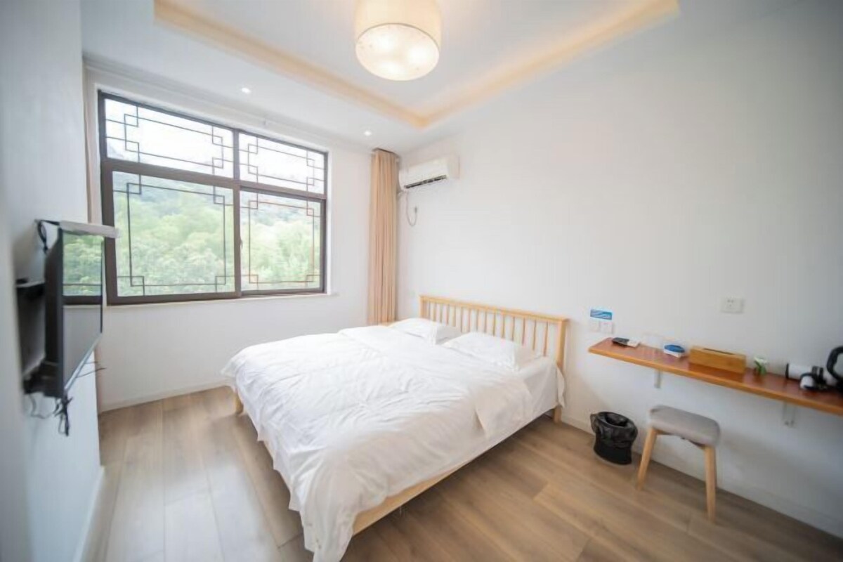 A bedroom with a bed, a TV, a desk, and a window with a view of greenery.