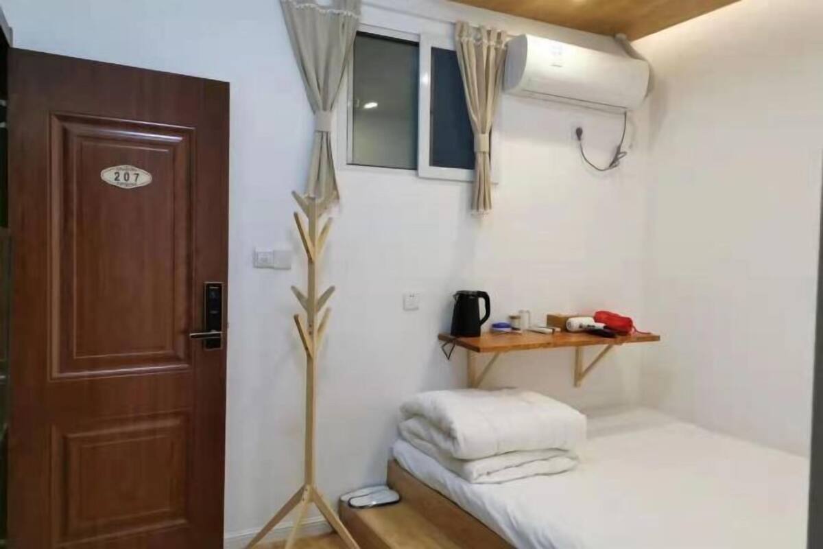 Une pièce avec une porte en bois portant le numéro « 207 », un lit avec une literie blanche, une petite étagère avec une bouilloire et d’autres objets, et une fenêtre avec des rideaux.