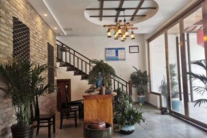Vestíbulo de hotel con escalera, mostrador de recepción de madera, plantas en macetas y ventanas amplias.