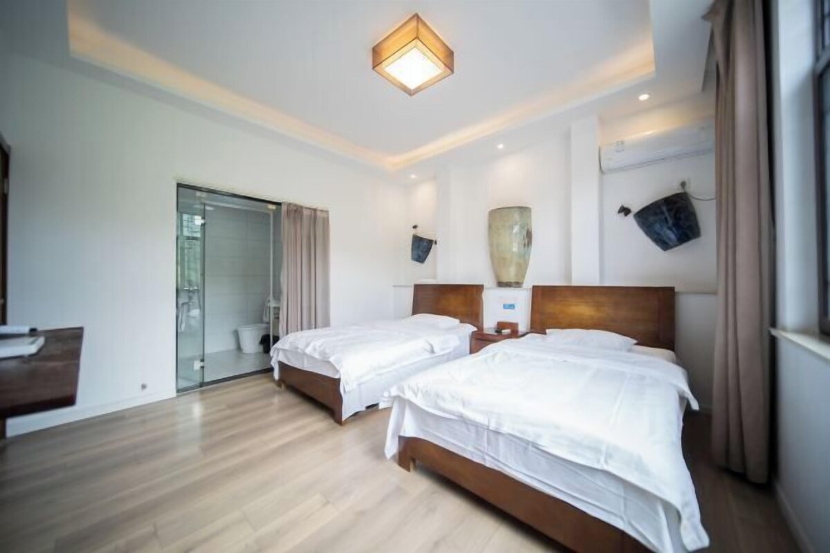 A hotel room with two beds, a wooden floor, a glass door leading to a bathroom, and a ceiling light fixture.