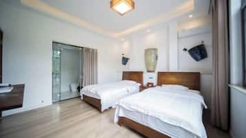 Habitación de hotel con dos camas, piso de madera, puerta de vidrio que da a un baño y una lámpara en el techo.