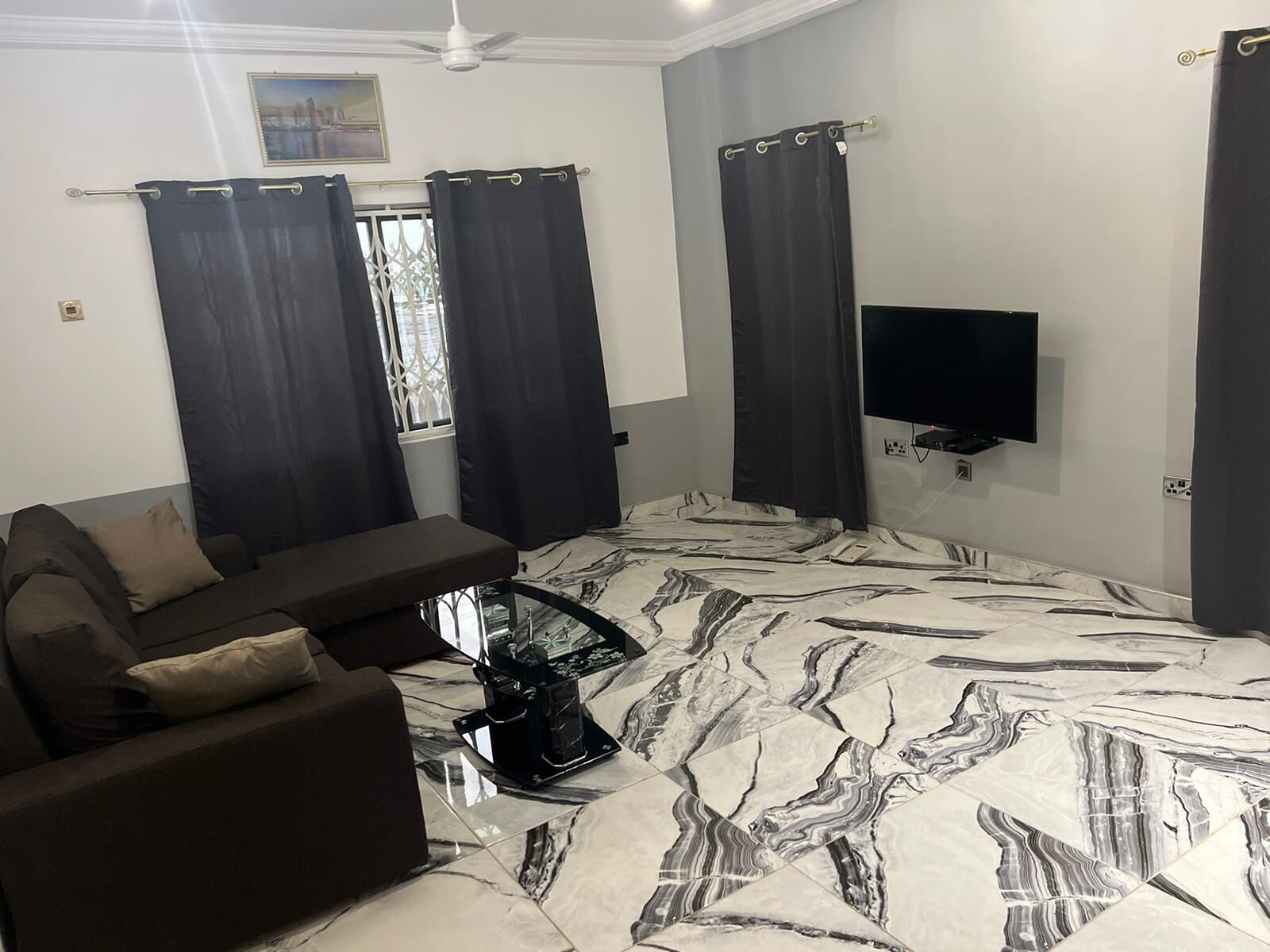 A living room with a sofa, a coffee table, a flat-screen TV mounted on the wall, and a window with curtains.
