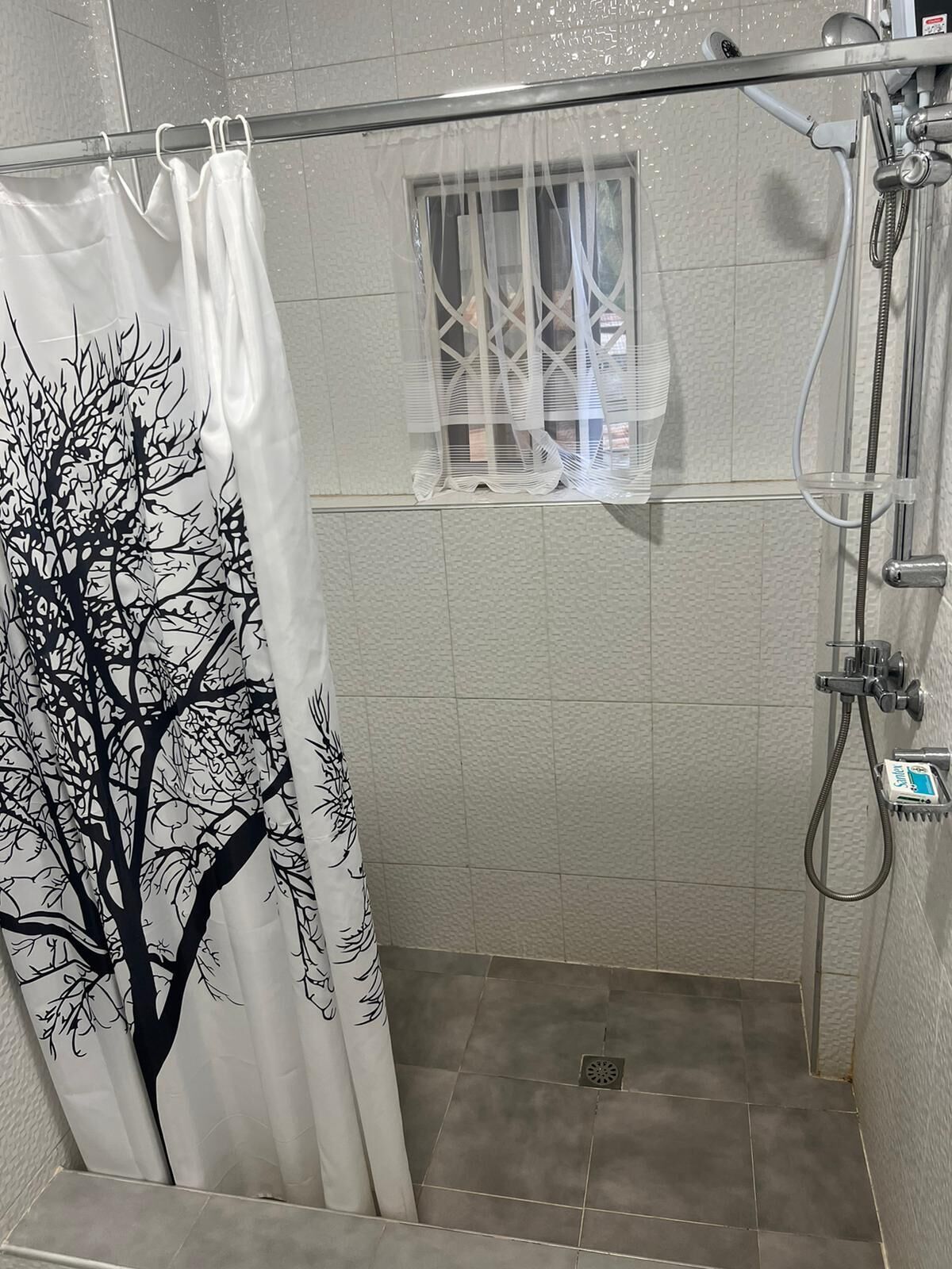 A shower area with a white shower curtain featuring a black tree design, a handheld showerhead, and a small window with bars.