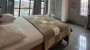 A spacious bedroom with a large bed, a ceiling fan, and a view of the city through large windows. - BAFFOUR APARTMENTS (Kumasi)