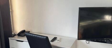 A hotel room with a white desk, a black office chair, a flat-screen TV, and a telephone.