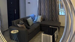 A living room with a sofa, a small side table, a radiator, and patterned curtains. - BAFFOUR APARTMENTS (Kumasi)