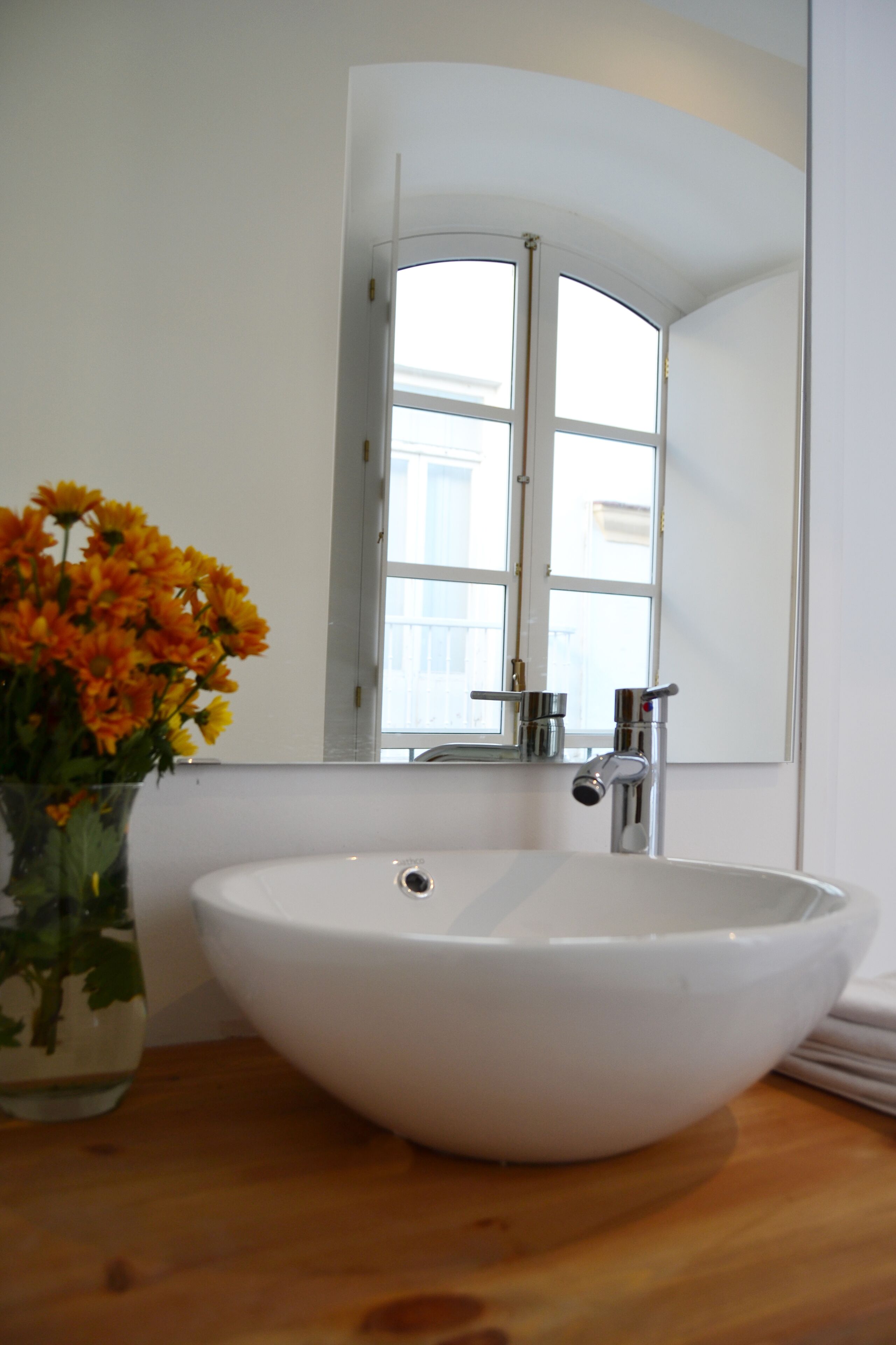 A white bathroom sink with a mirror above it, a window, and a vase of flowers on the counter.