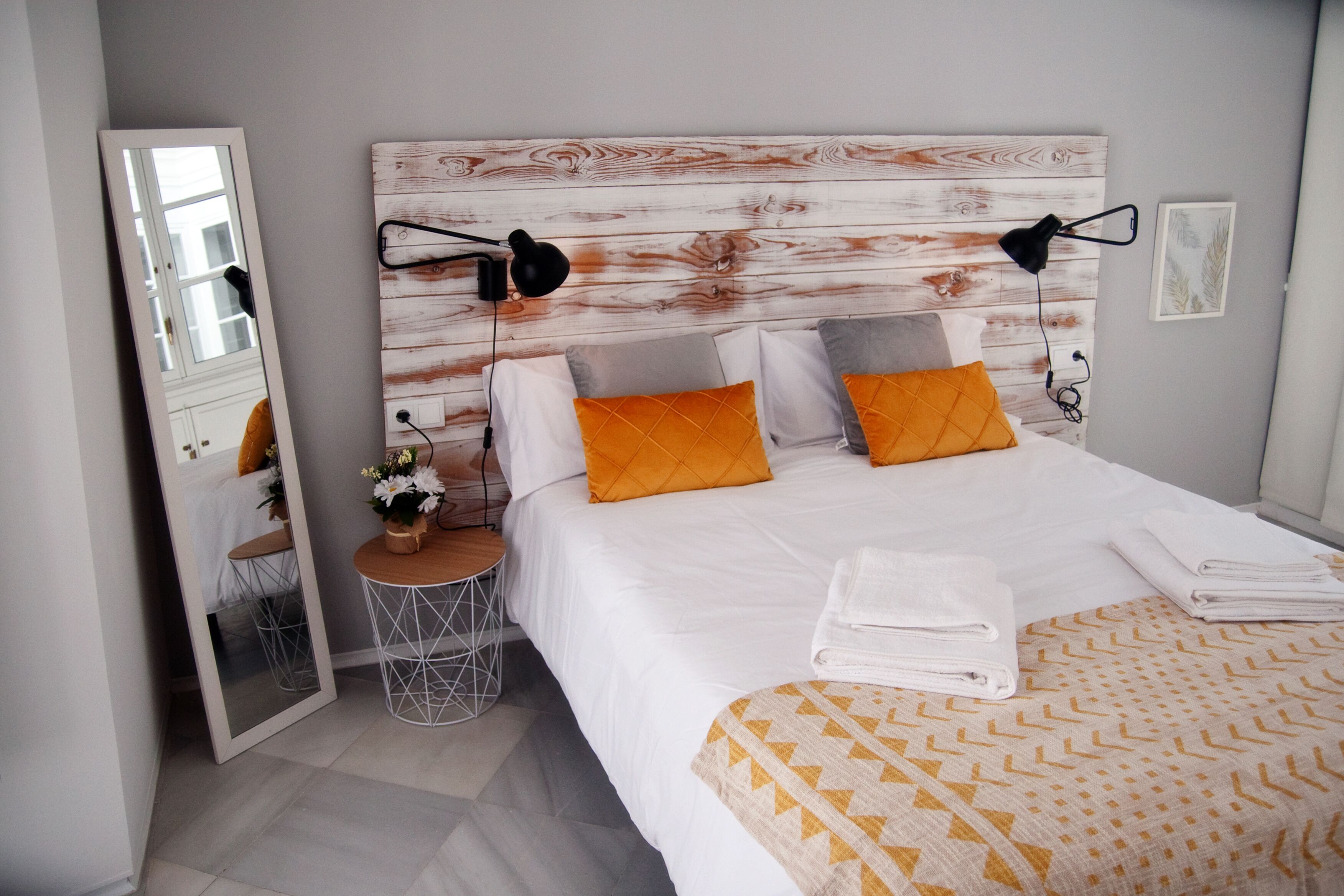 A neatly arranged bedroom with a large bed, a bedside table with a lamp, a mirror, and a small round table with a vase of flowers.