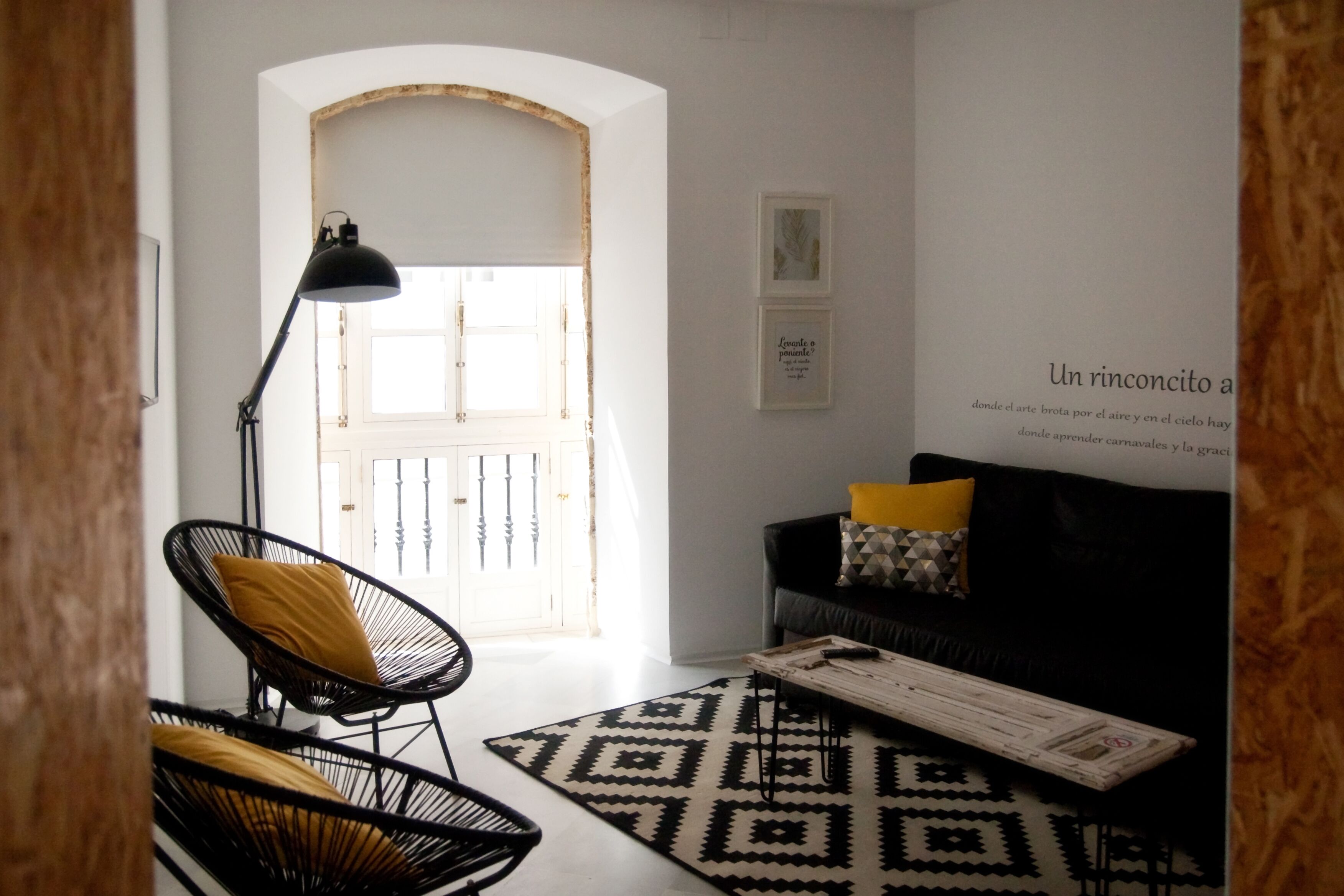 A modern living room with a black and white patterned rug, a black chair with a yellow cushion, a black sofa with a geometric pillow, and a black floor lamp.