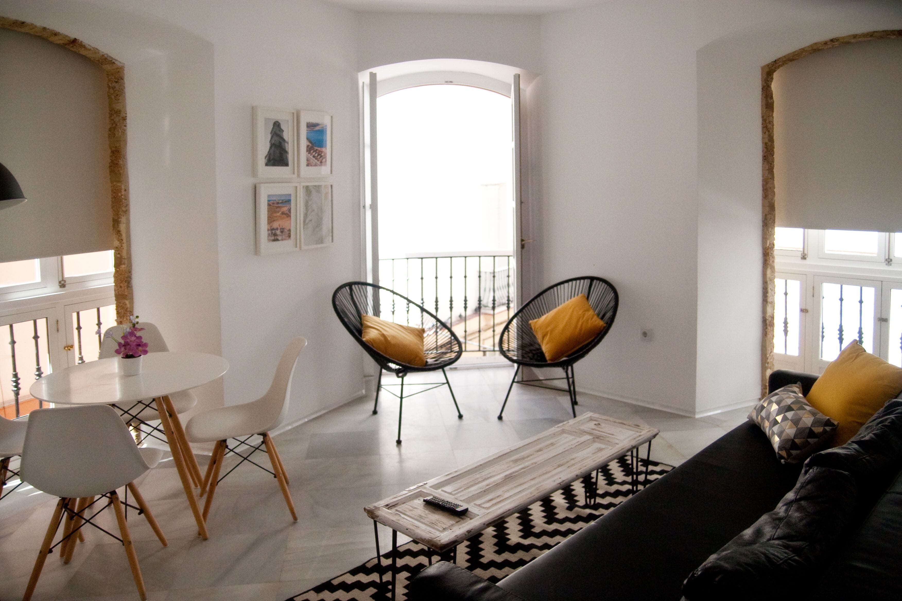 Un salón con un sofá de cuero negro, dos sillas metálicas negras con cojines amarillos, una mesa redonda blanca con un florero y un tapete de diseño blanco y negro.