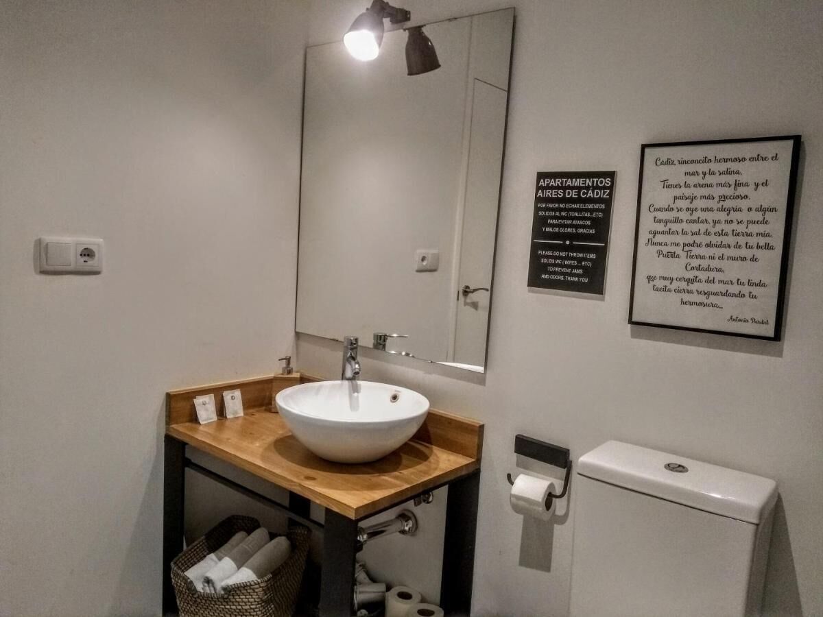 Un baño con lavamanos blanco, encimera de madera y un espejo encima. Hay dos citas enmarcadas en la pared, un inodoro y un cubo de basura.