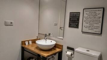 Un baño con lavamanos blanco, encimera de madera y un espejo encima. Hay dos citas enmarcadas en la pared, un inodoro y un cubo de basura.