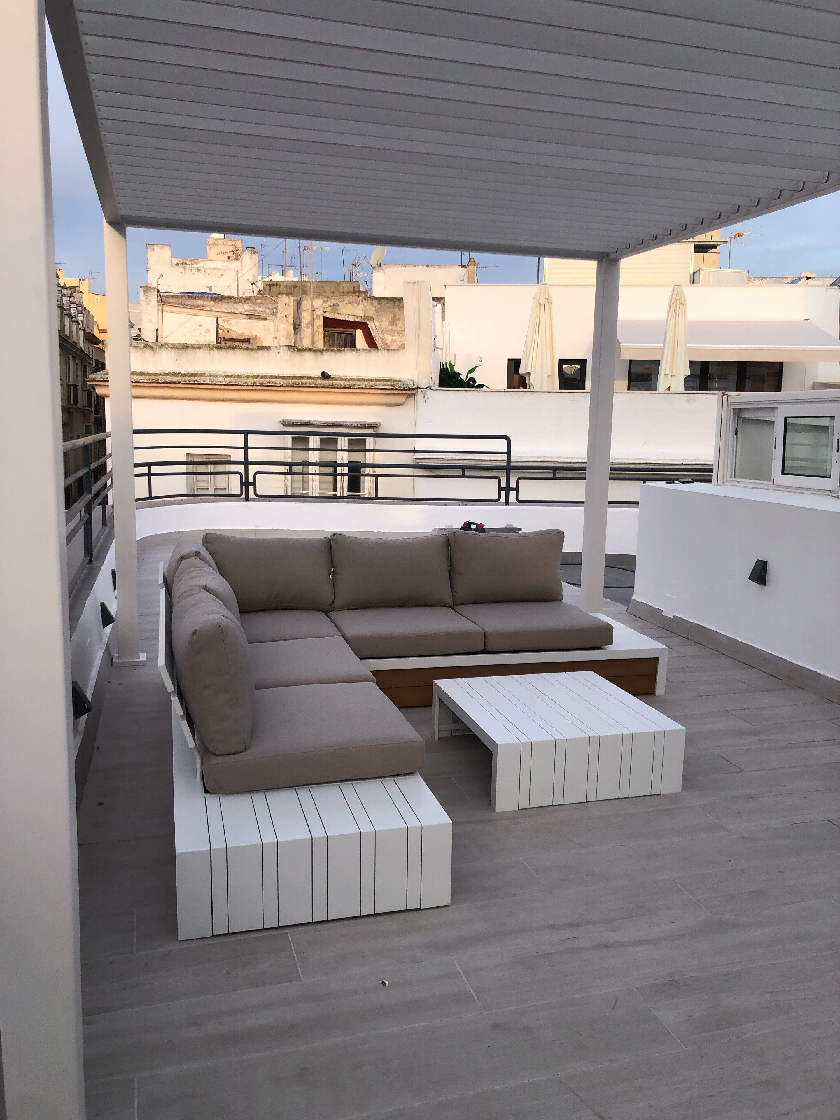 Een dakterras met een beige modulaire bank, een witte salontafel en een pergola erboven.
