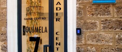 A building entrance with a glass door displaying 'GADIR CENTRO' and 'COLUMELA 7'. A blue sign with 'AT' and 'EDIFICIO/COMPLEJO CIUDAD' is visible.