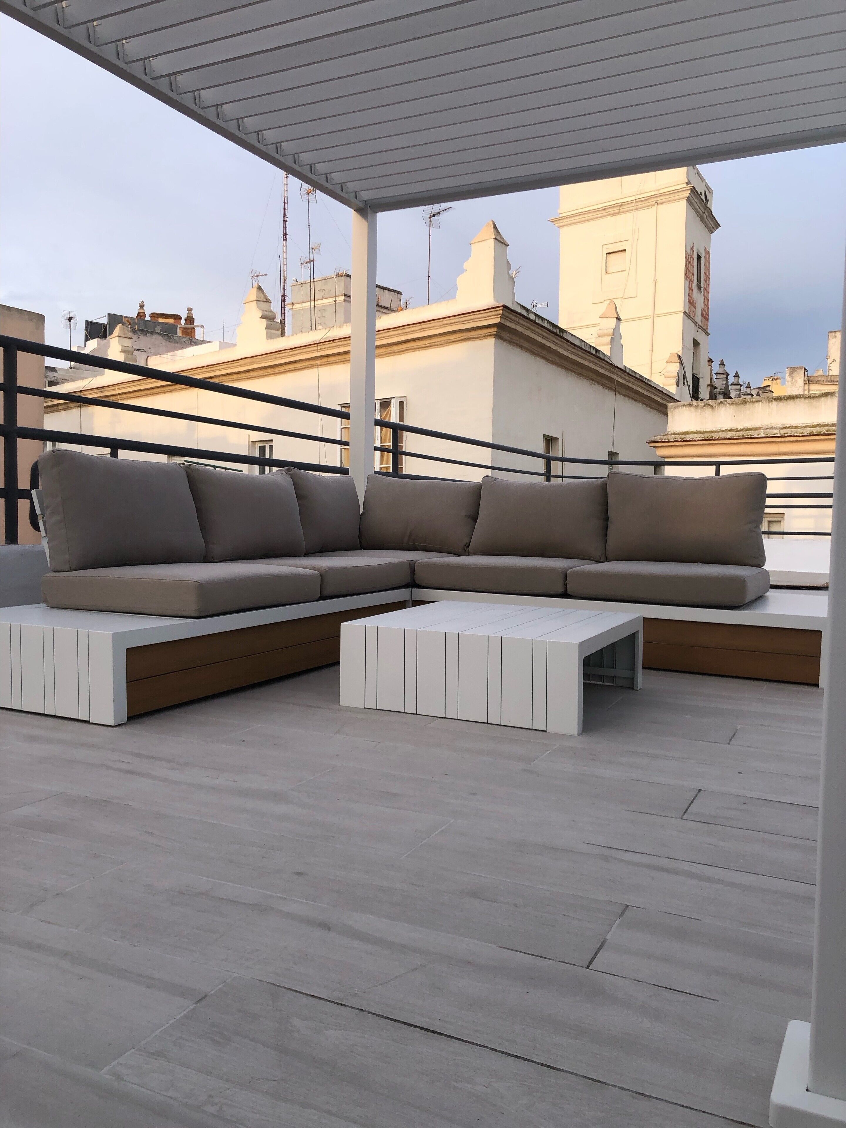 A modern outdoor seating area with a sectional sofa and a coffee table, set on a rooftop terrace with a view of surrounding buildings.