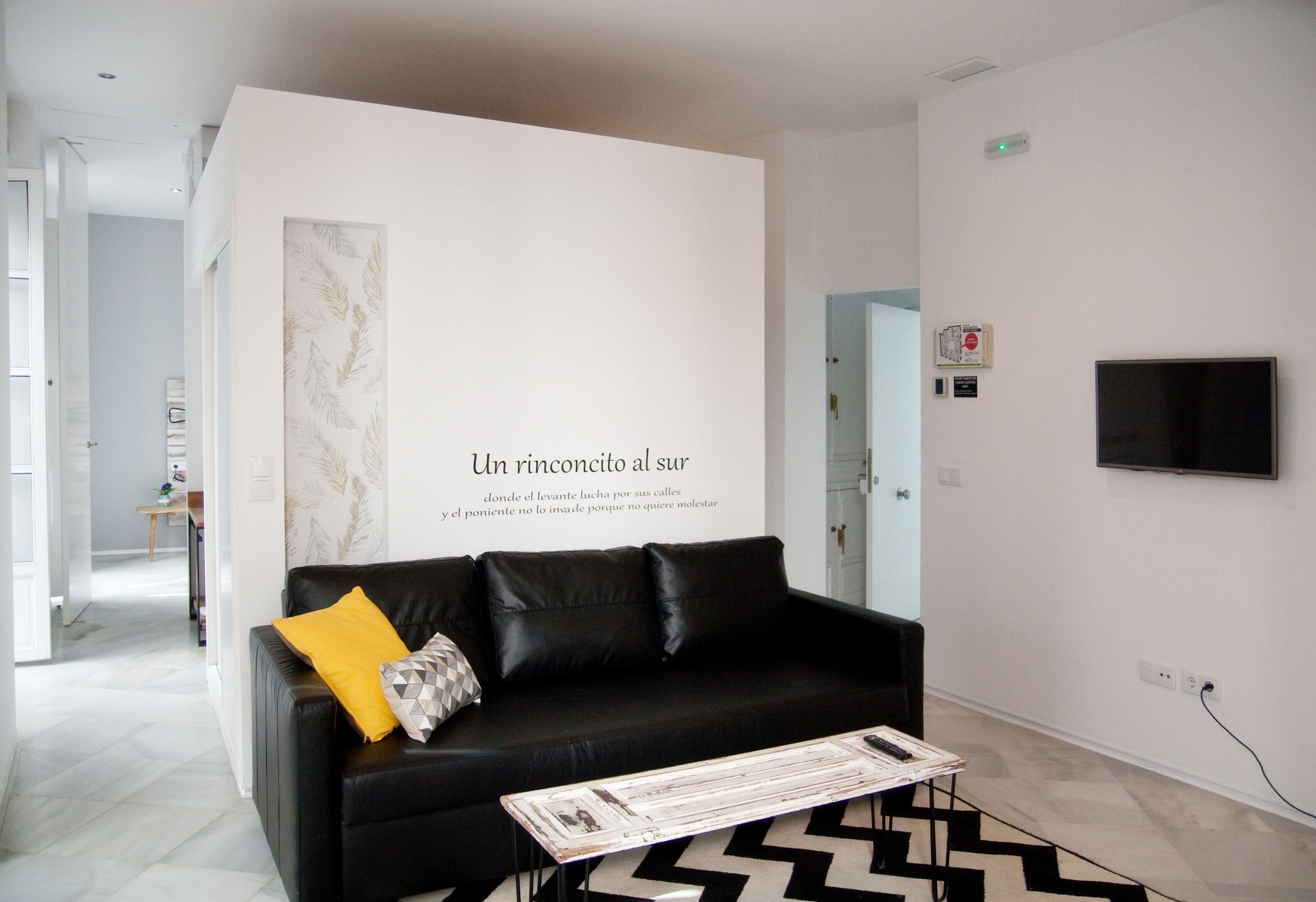 A modern living room with a black leather sofa, a yellow pillow, and a magazine on a coffee table. A large wall-mounted TV and a framed quote are visible.