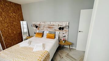 A modern bedroom with a wooden headboard, a bed with white linens and yellow accents, a bedside table with a lamp, and a small round table with a flower vase.