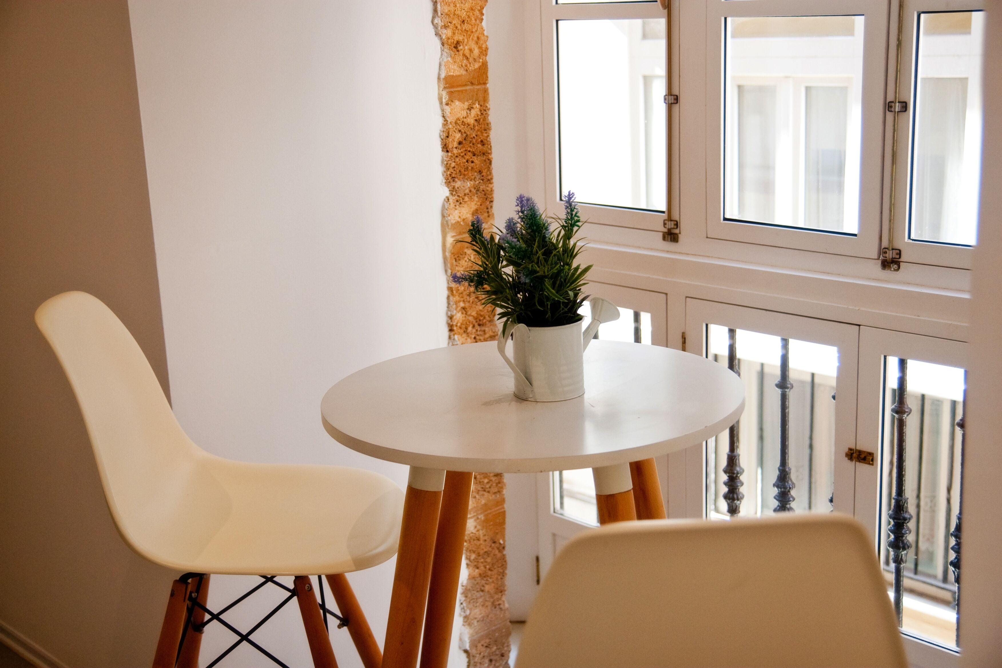A small round table with two white chairs, a potted plant, and a window with a view of a building.