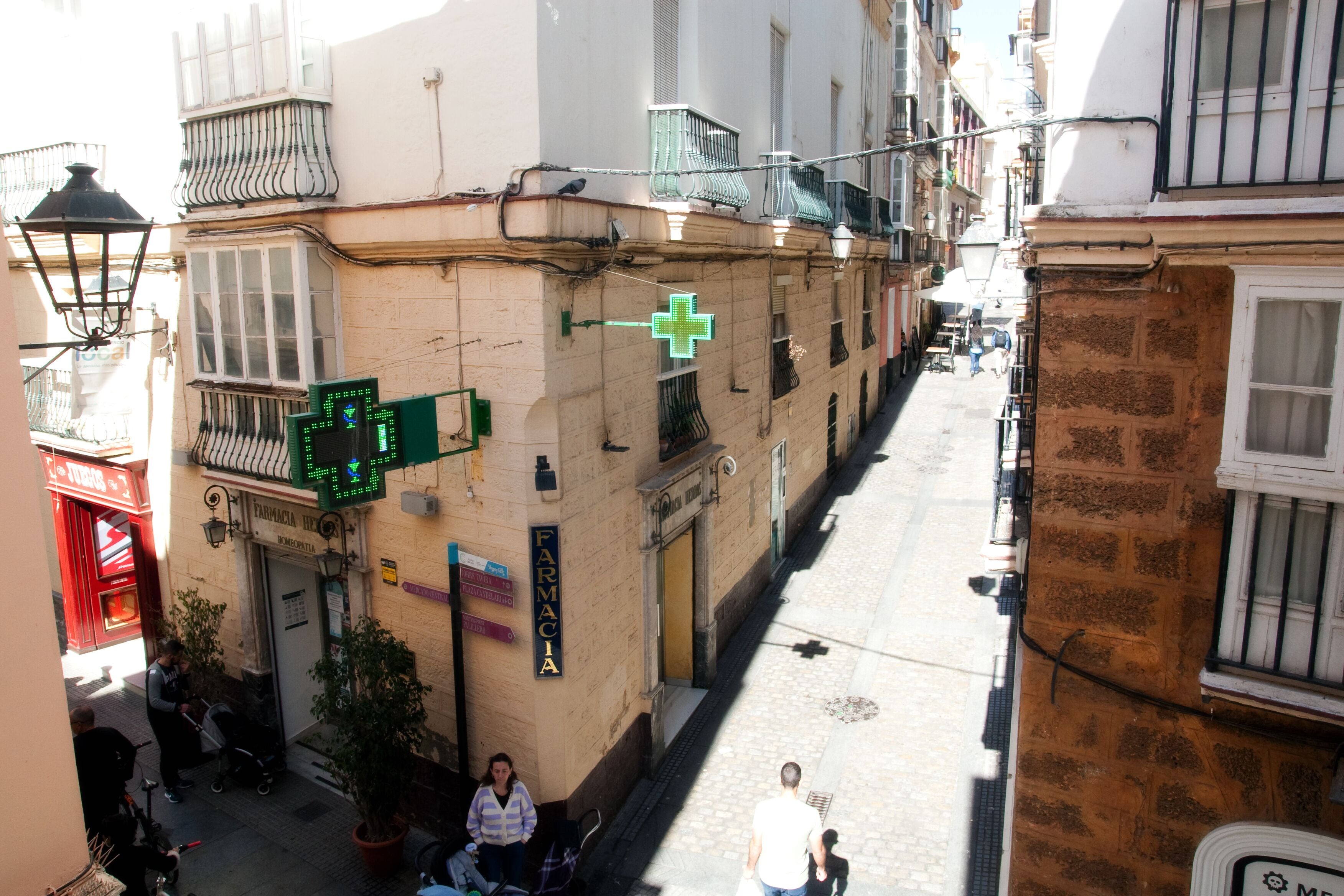 Una calle estrecha flanqueada por edificios, una farmacia con un cartel de cruz verde y un escaparate rojo.