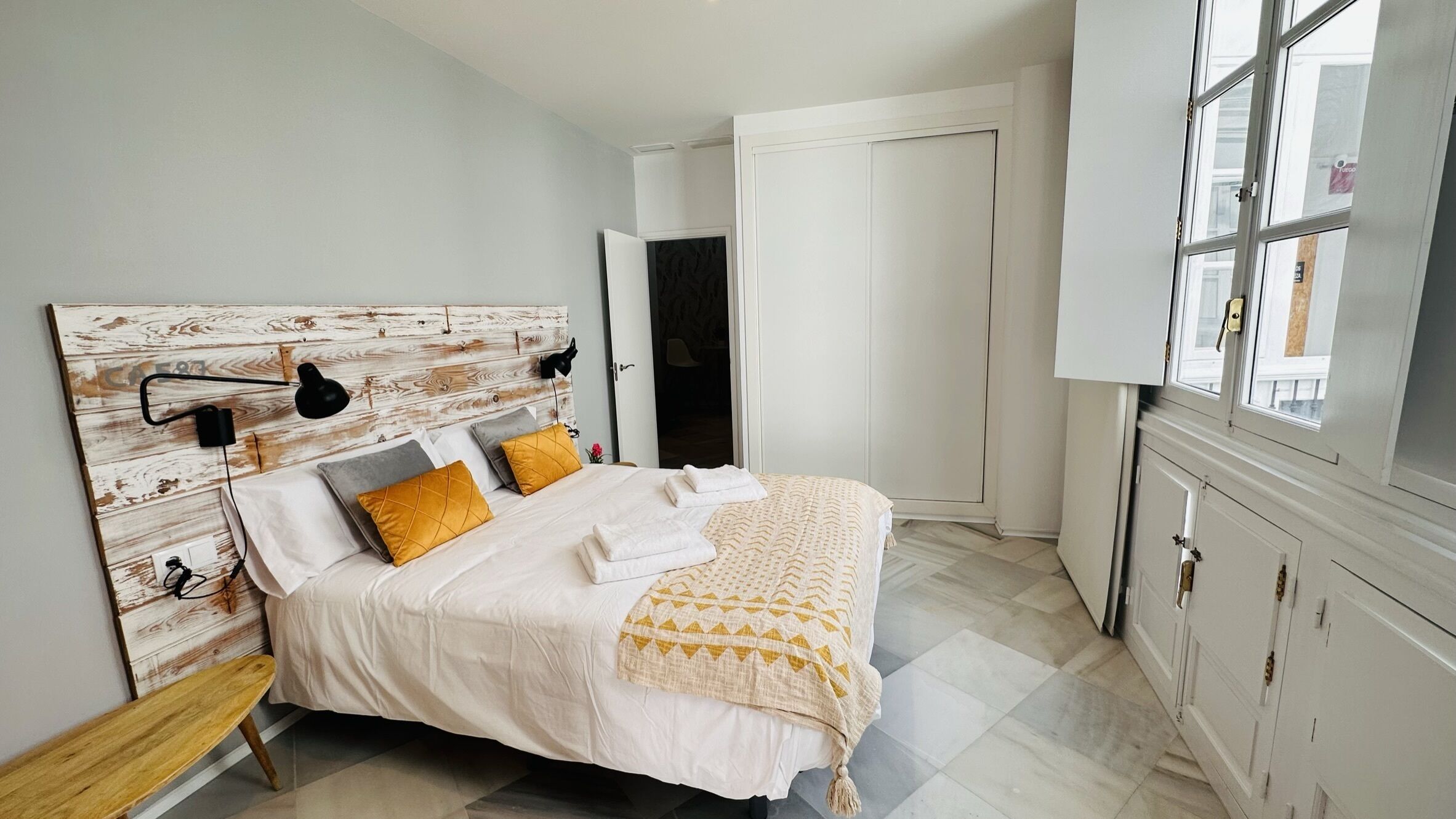 A bedroom with a wooden headboard, a bed with white bedding and yellow pillows, a bench, and a window with a view.
