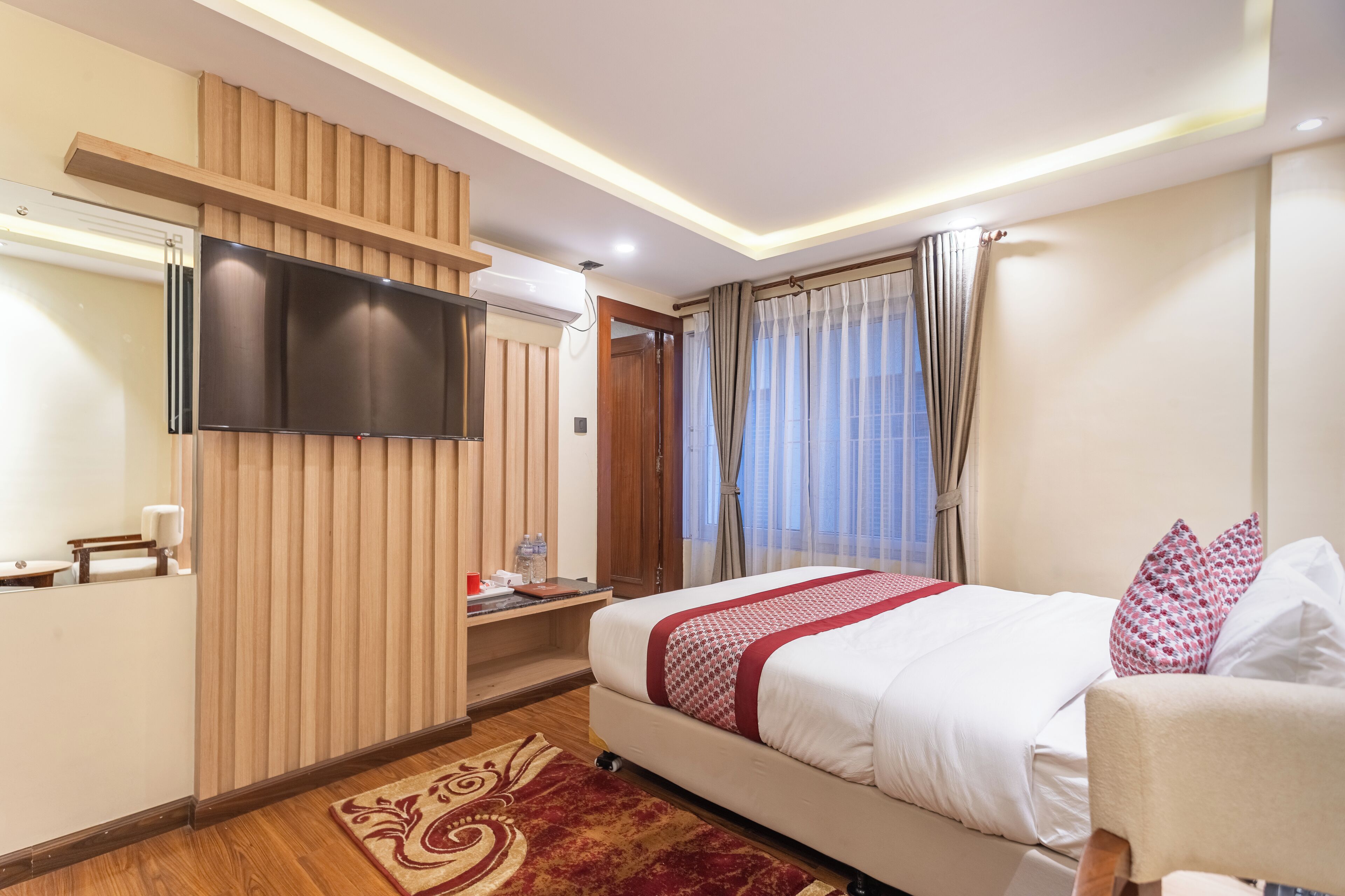 A hotel room with a bed, a TV mounted on the wall, a wooden panel design, a window with curtains, and a small table with a water bottle.