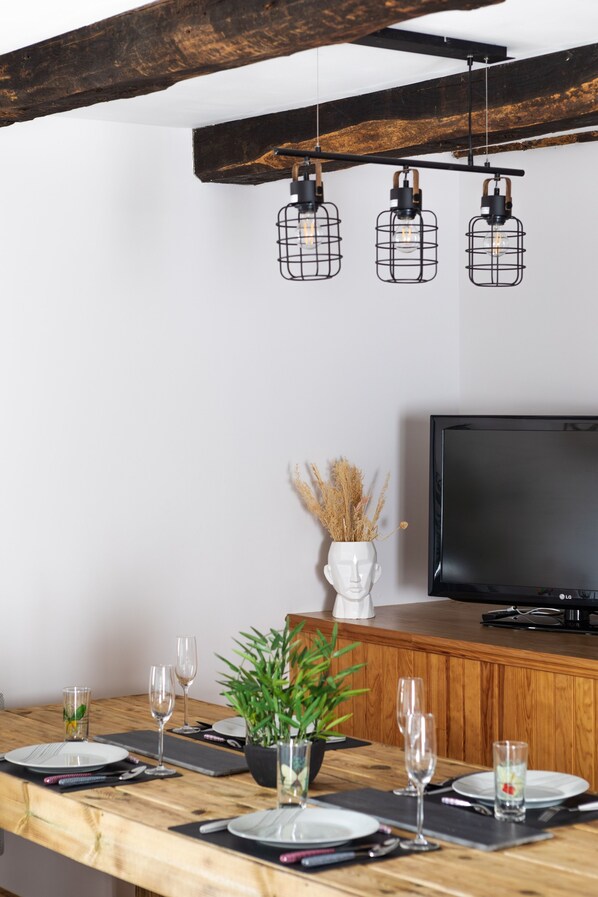 Un comedor moderno con una mesa de madera dispuesta para cuatro personas, un televisor y lámparas colgantes.