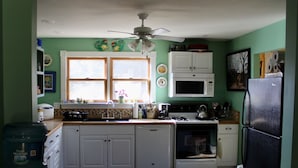 A kitchen with white cabinets, a microwave, stove, and a refrigerator. There are two windows with curtains, a ceiling fan, and a rug on the floor. - Charming 1920s Hillside Home Overlooking Valley and Rails-To-Trails (Rimersburg)