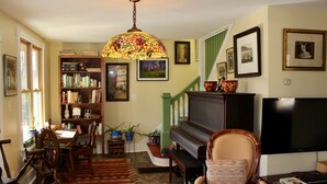 A cozy living room with a piano, a bookshelf, a television, and a decorative stained glass lamp. - Charming 1920s Hillside Home Overlooking Valley and Rails-To-Trails (Rimersburg)