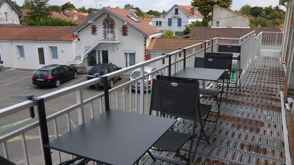 A balcony with metal railings, a table, and chairs. There are parked cars in the background.