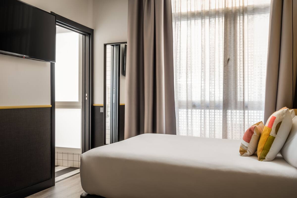 Quarto de hotel com cama, televisão, guarda-roupa e janela com cortinas.