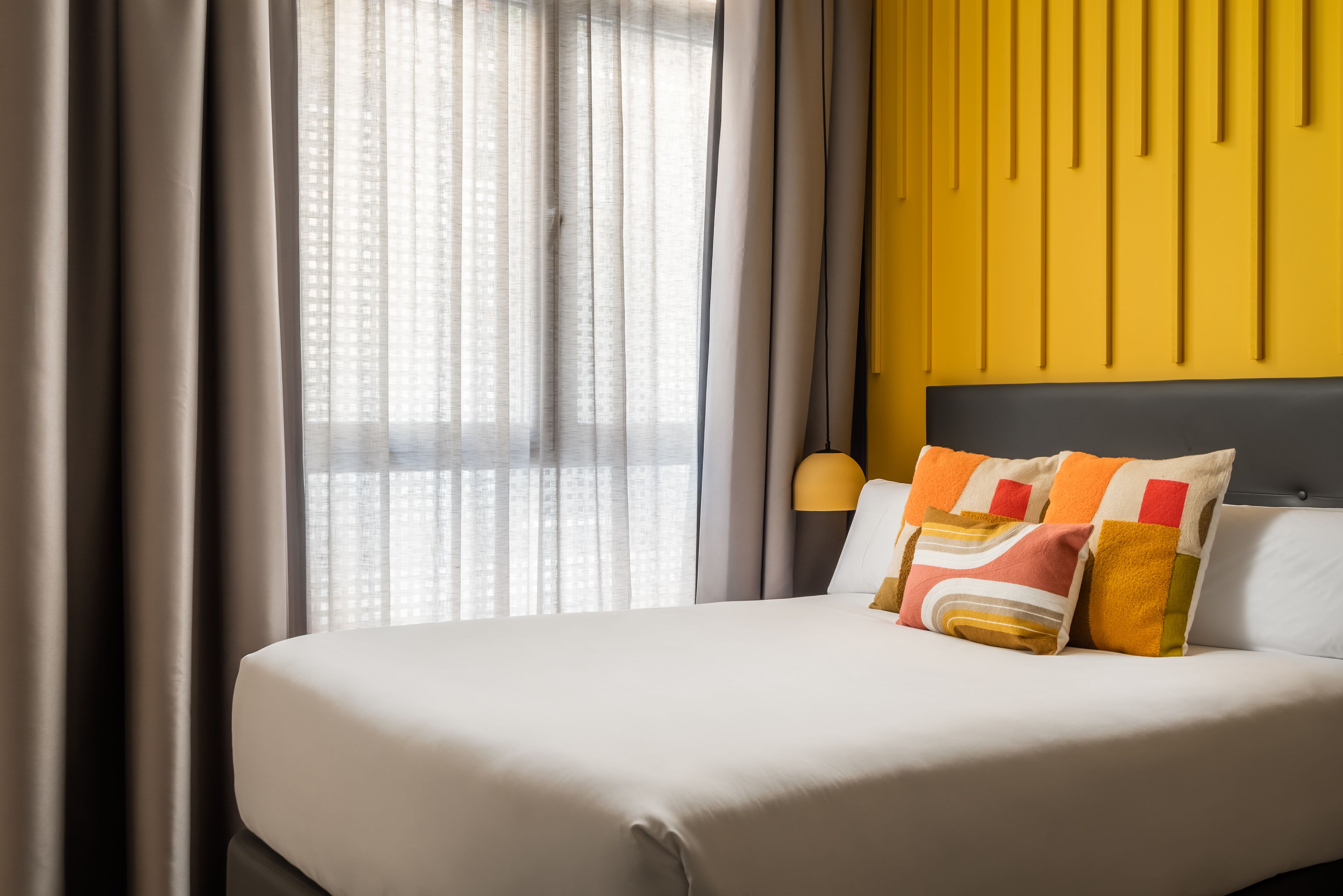 A neatly made bed with a white mattress and colorful pillows, a yellow accent wall, and a window with sheer curtains.