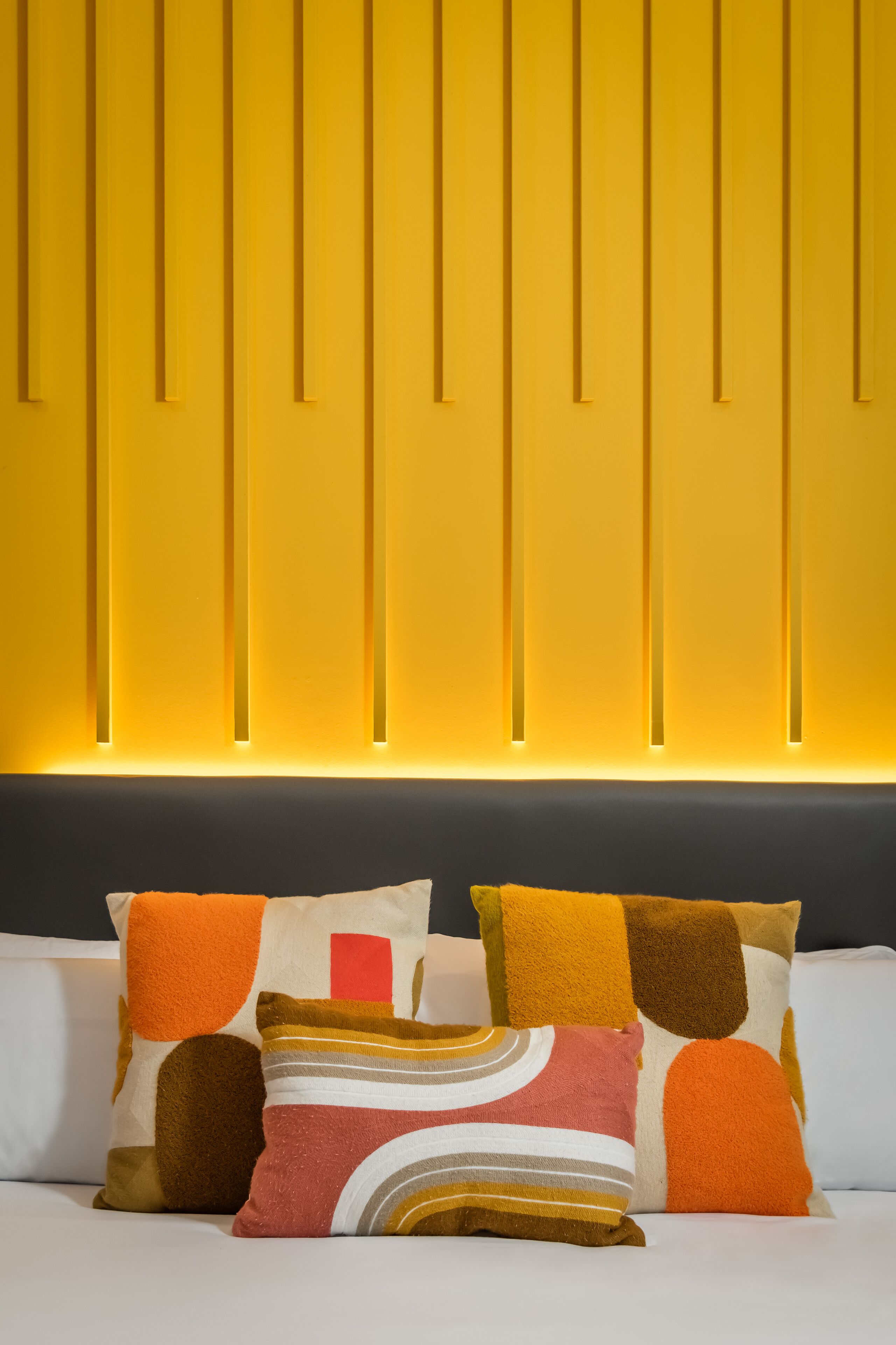 Une chambre d’hôtel au design moderne, avec un mur jaune orné de lignes verticales et une tête de lit sombre. Le lit est agrémenté de trois oreillers, chacun ayant des motifs et des couleurs différents.