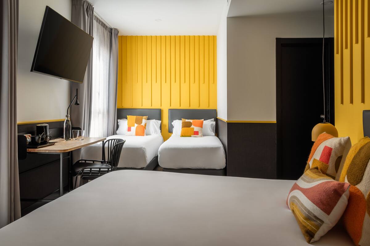 Quarto de hotel com duas camas, uma escrivaninha com cadeira, uma TV de tela plana na parede e uma janela com cortinas.