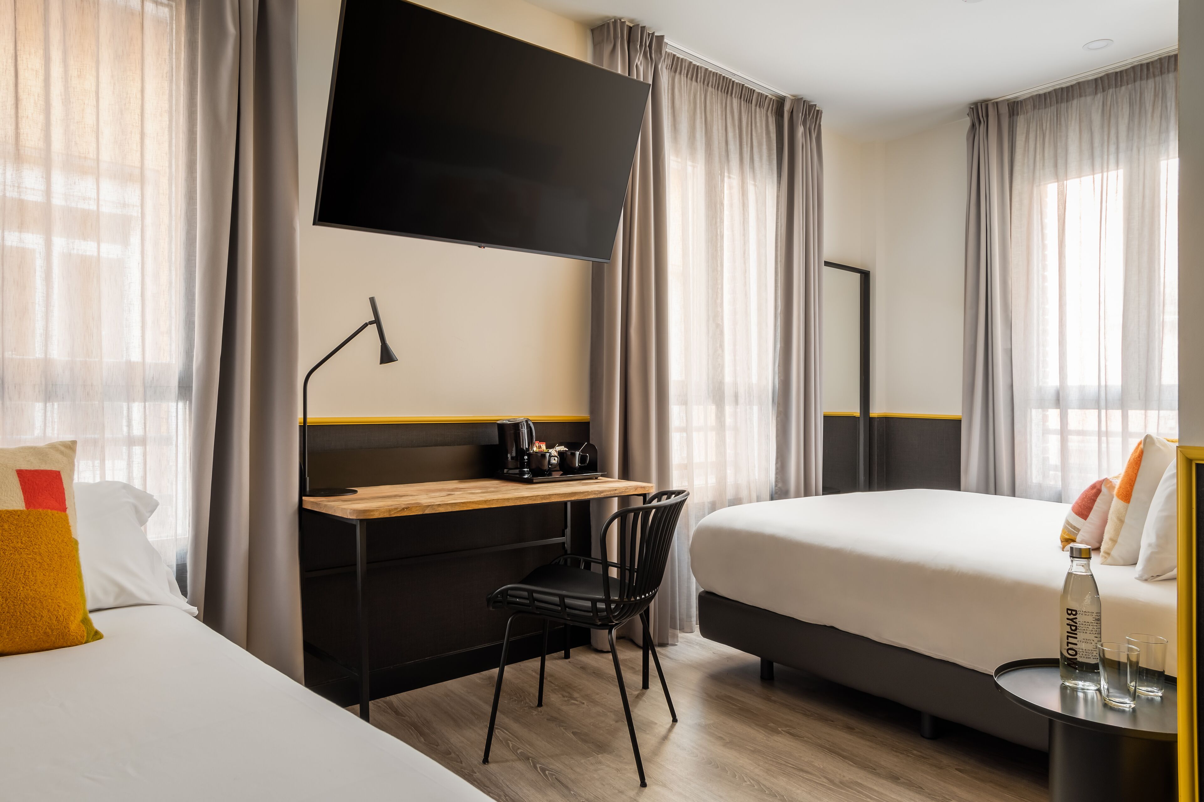 A hotel room with a bed, a desk with a chair, a flat-screen TV, and a bedside table with glasses and a bottle.