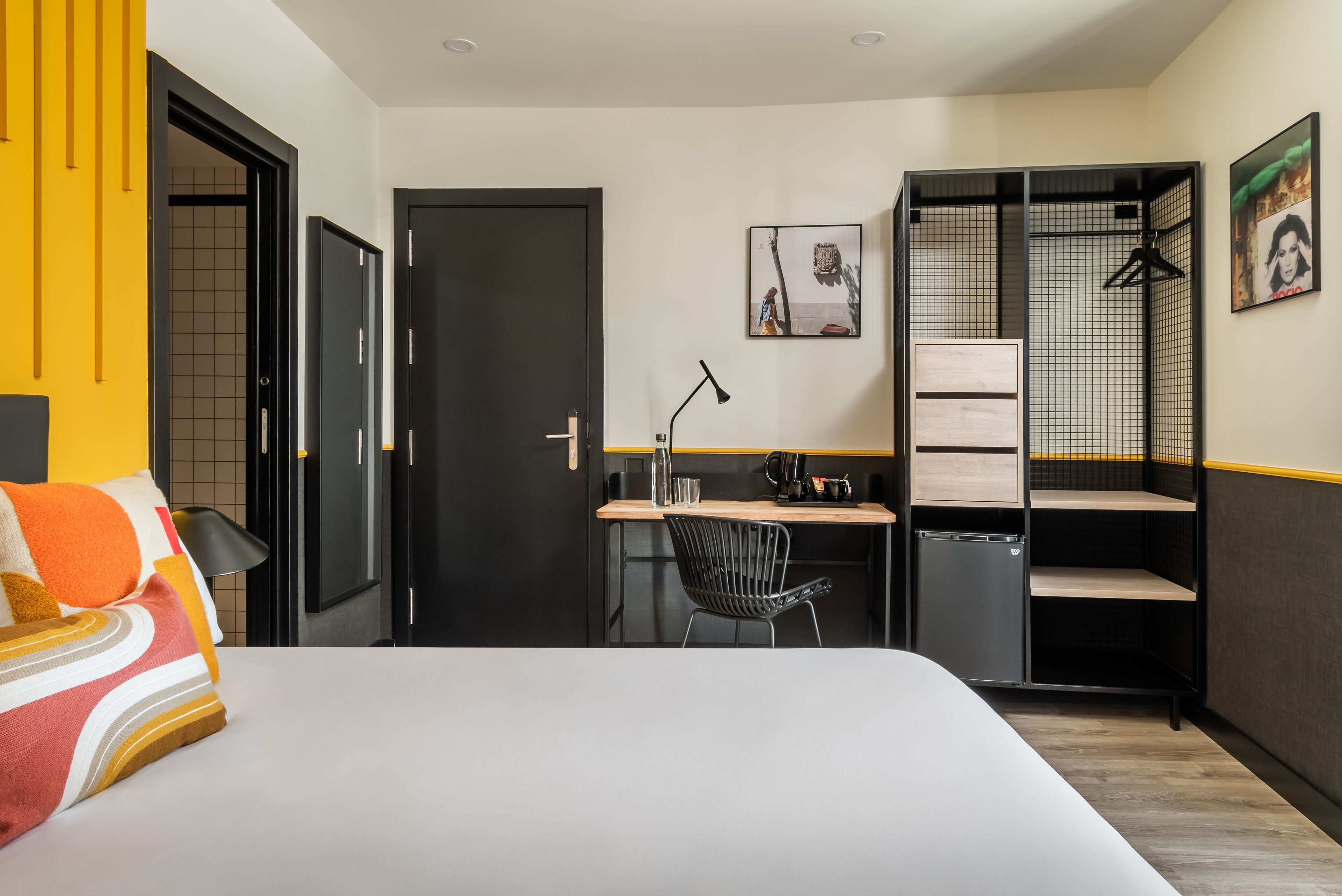 A hotel room with a bed, a desk, a chair, a small fridge, and a wardrobe.