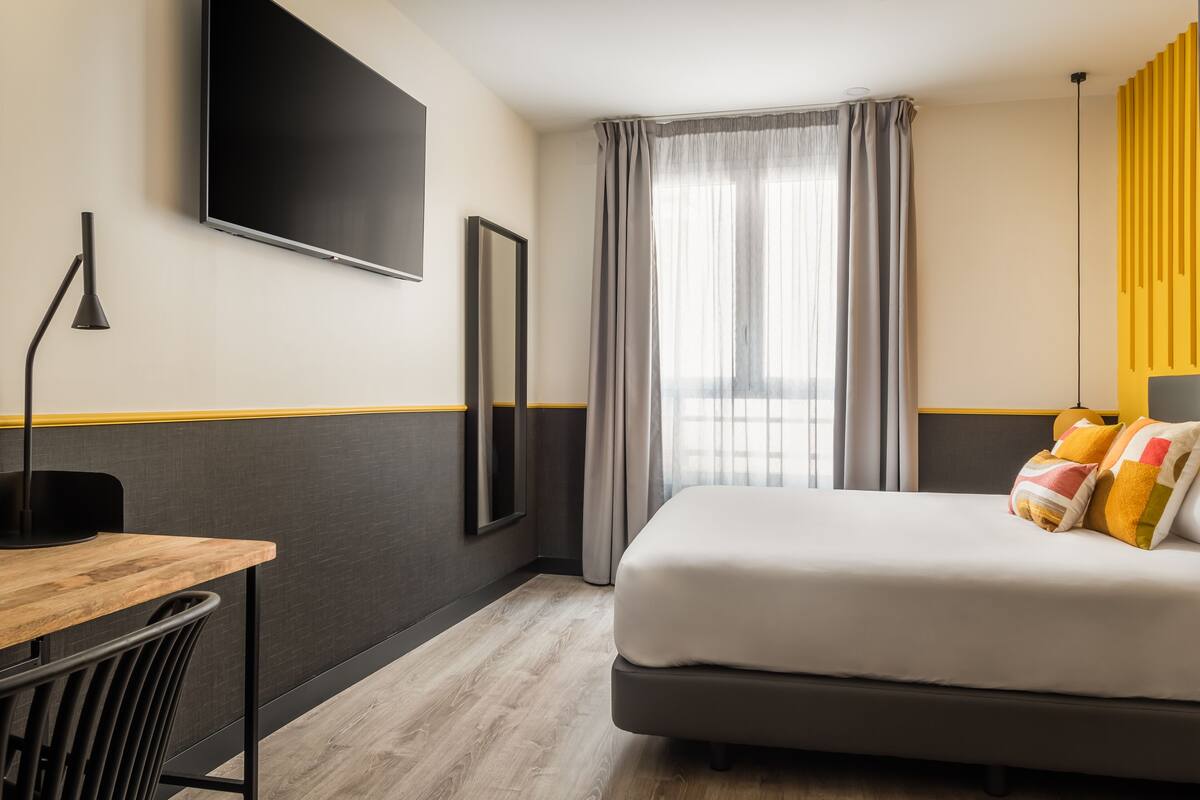 Quarto de hotel com cama, escrivaninha, cadeira, televisão e janela com cortinas.