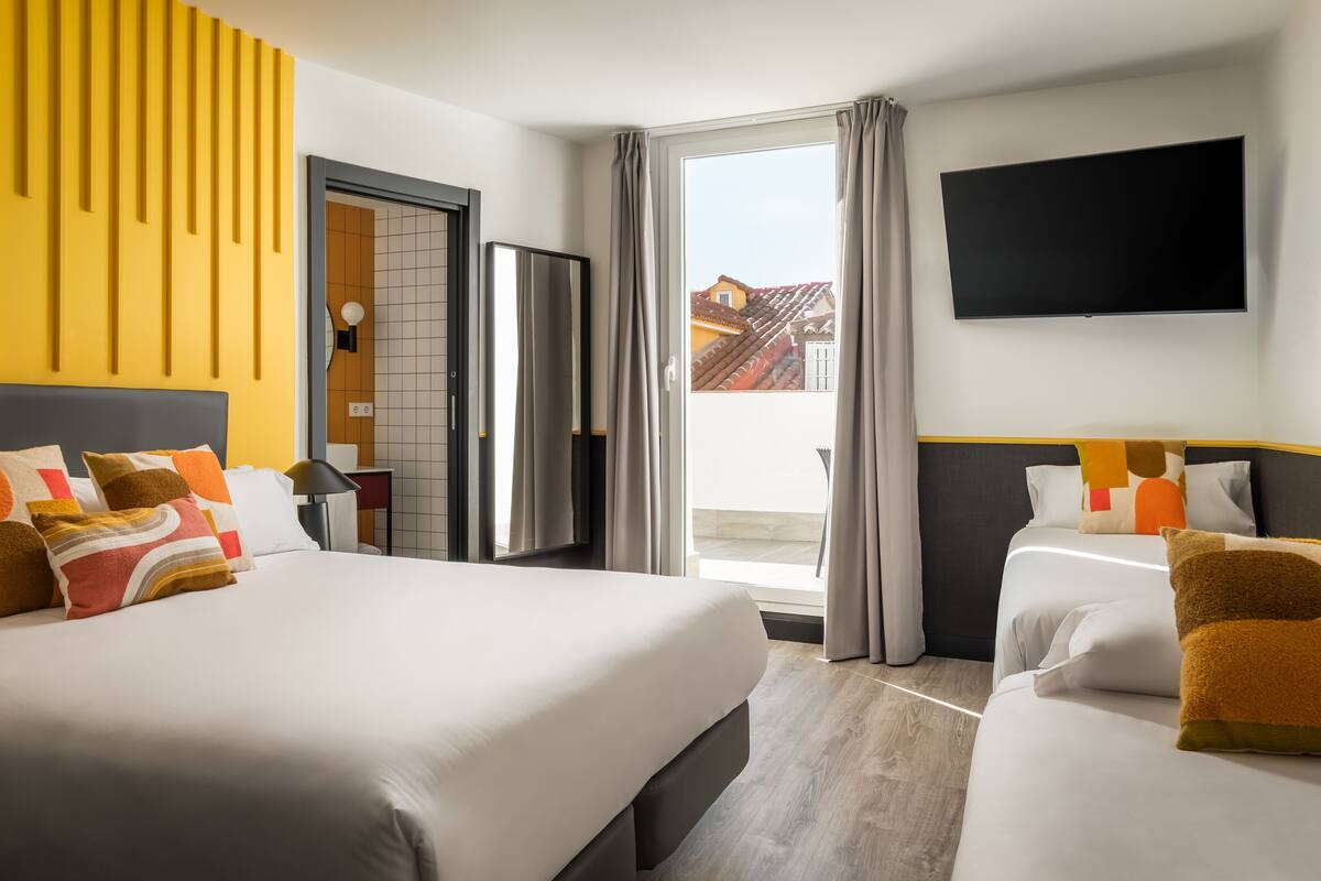 Quarto de hotel com duas camas, televisão, porta de correr e vista para edifícios.
