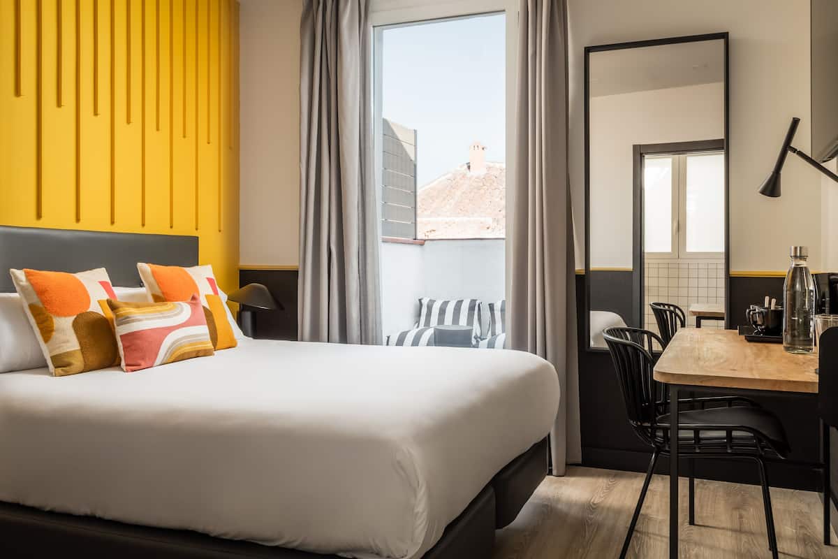 Quarto de hotel com cama, mesa de jantar, cadeiras, abajur e vista da cidade pela janela.
