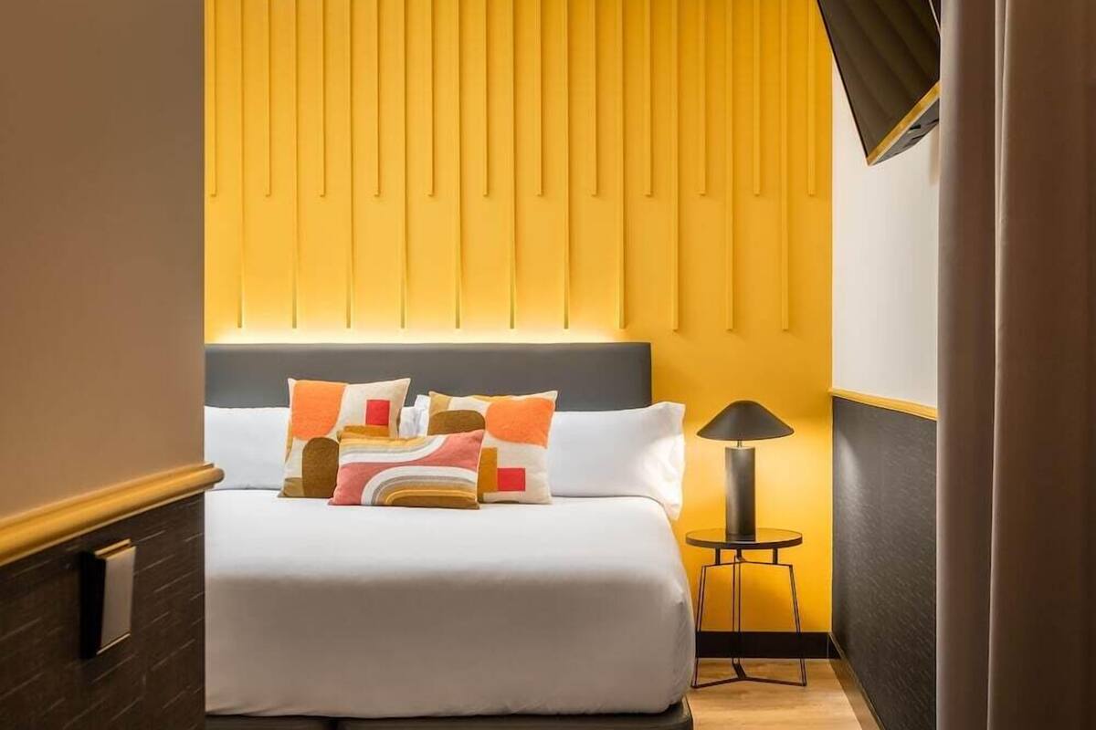 Quarto de hotel com cama, almofadas, mesa de cabeceira com abajur e uma parede com painéis verticais.