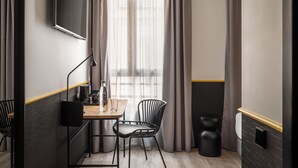 Una habitación de hotel moderna con un escritorio de madera, dos sillas, una lámpara de pie negra, un ventanal con cortinas translúcidas y un cubo de basura negro.
