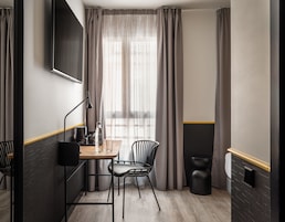 A modern hotel room with a wooden desk, two chairs, a black floor lamp, a large window with sheer curtains, and a black trash bin.