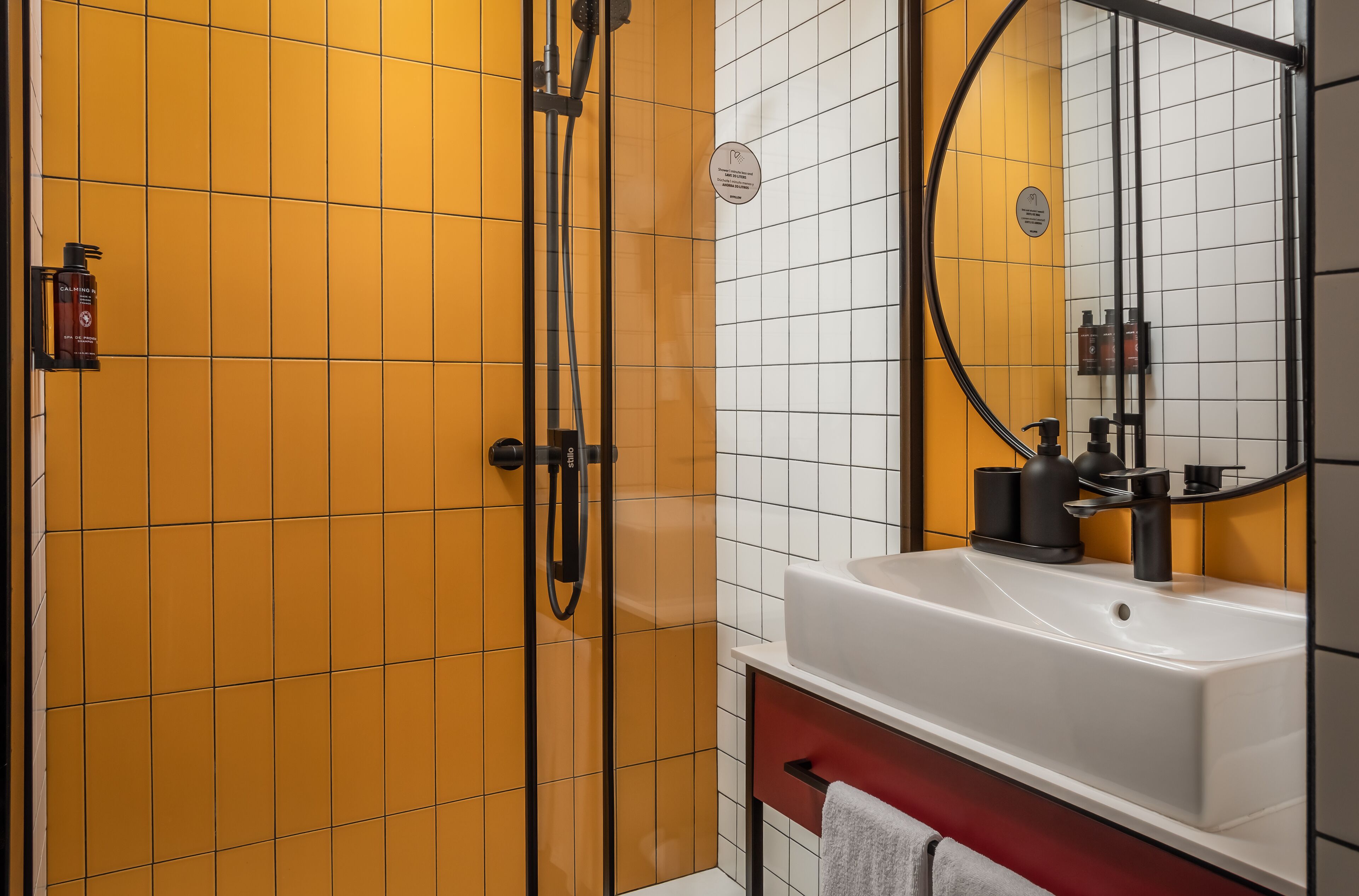 Un baño moderno con una cabina de ducha con azulejos amarillos, un lavabo blanco con espejo y un tocador rojo.