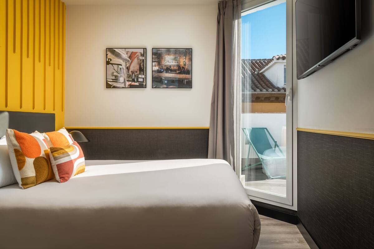Quarto de hotel com cama, duas obras de arte penduradas na parede, uma janela ampla e televisão na parede.