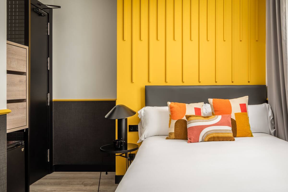 Quarto de hotel com cama, mesa de cabeceira, luminária e guarda-roupa.