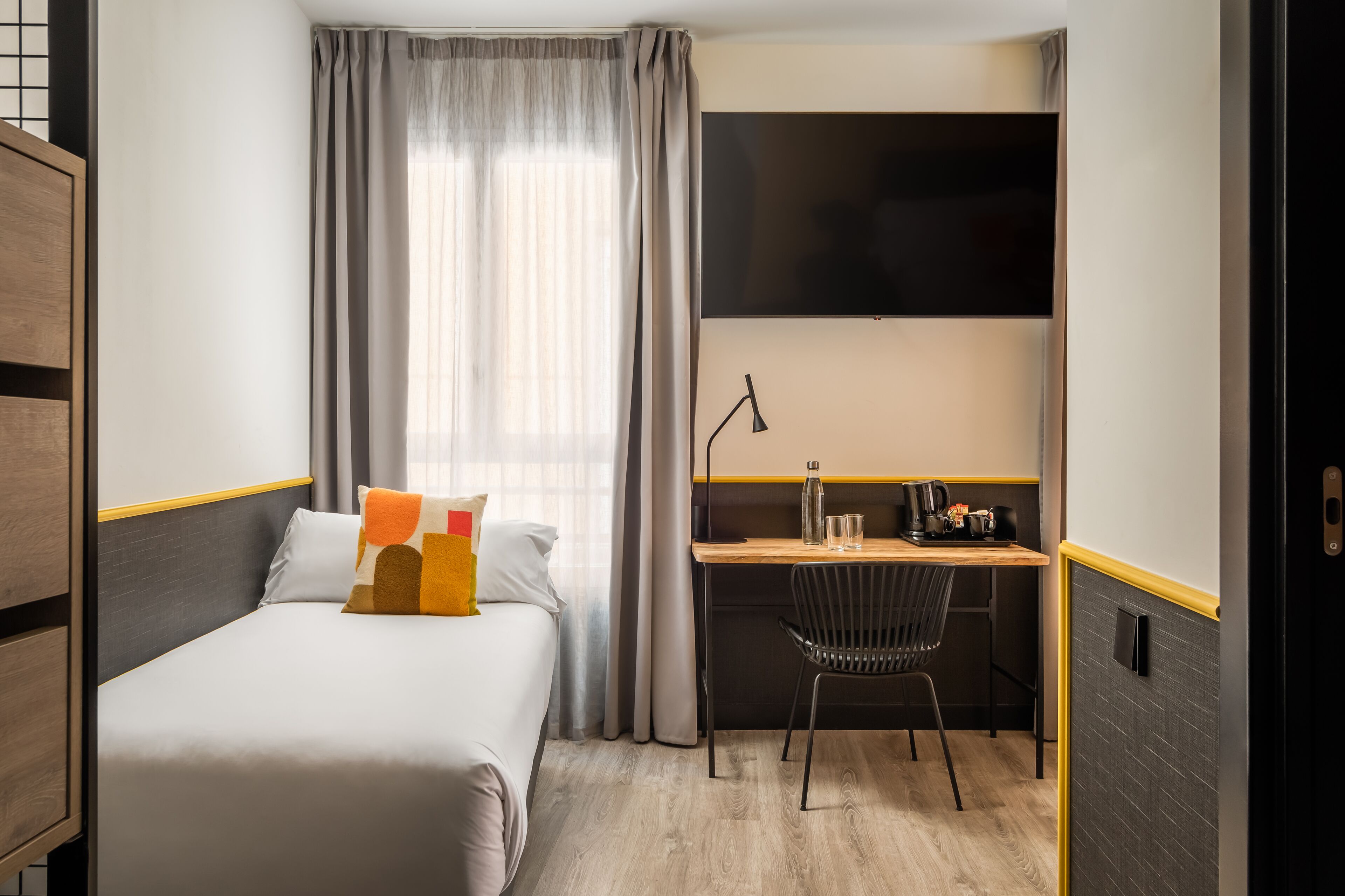 A compact hotel room with a single bed, a desk with a chair, a flat-screen TV, and a window with curtains.