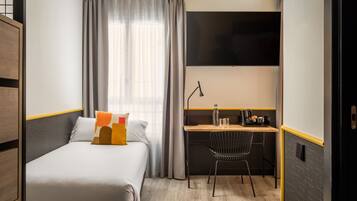 A compact hotel room with a single bed, a desk with a chair, a flat-screen TV, and a window with curtains.
