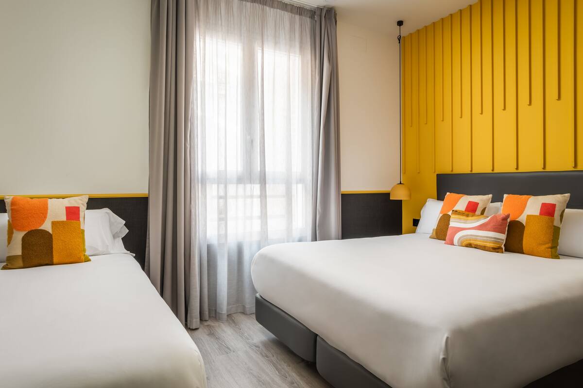 Quarto de hotel com duas camas, uma janela grande com cortinas leves, uma parede de destaque amarela e uma luminária moderna.
